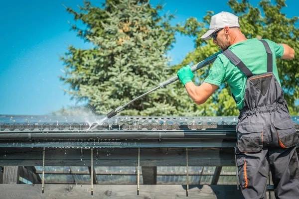 A worker in gloves and overalls rinsing out a roof gutter with a pressure washer wand, with evergreen trees behind